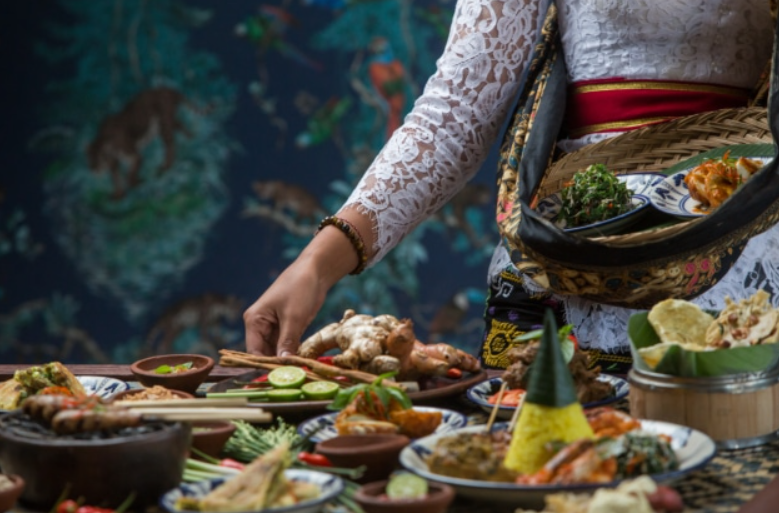 A woman in traditional Balinese attire serves a variety of simple, tasty Balinese food on a table, showcasing authentic local cuisine in a relaxed setting.