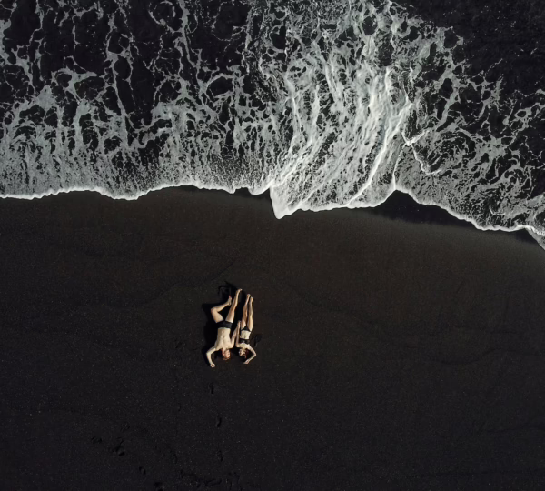 Drone view of relaxing hugging couple in dark swimwear chilling on black sand beach near splashing waving sea in daytime