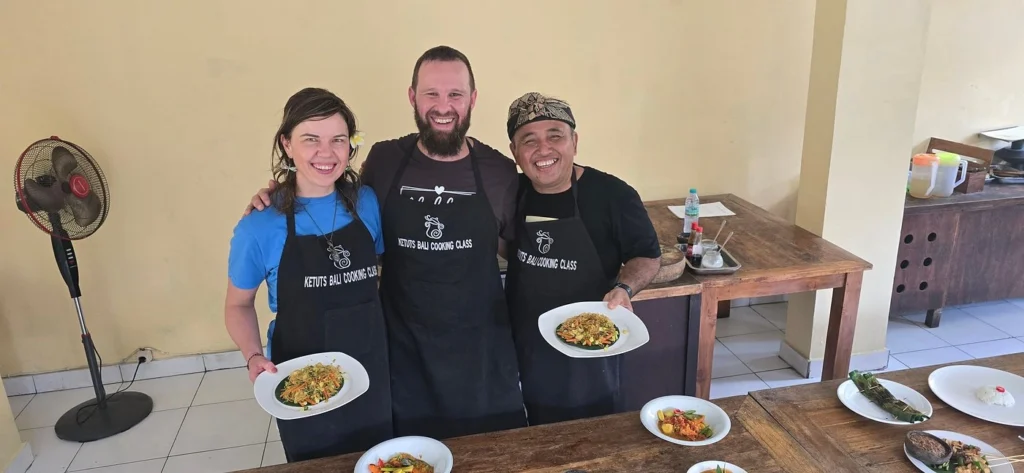 Chef Ketut teaches students traditional Balinese cooking techniques in a friendly class. This image features Ketut's Bali Cooking Class in Ubud. Chef Ketut is guiding students through traditional Balinese recipes, showcasing his popular hands-on teaching style.
