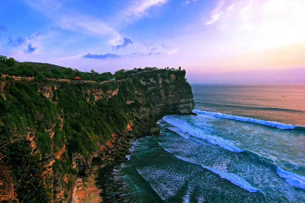 A surfer catching a wave below a dramatic cliff at Uluwatu, with the iconic Uluwatu Temple visible in the distance, emphasizing the area's natural beauty and surf culture.