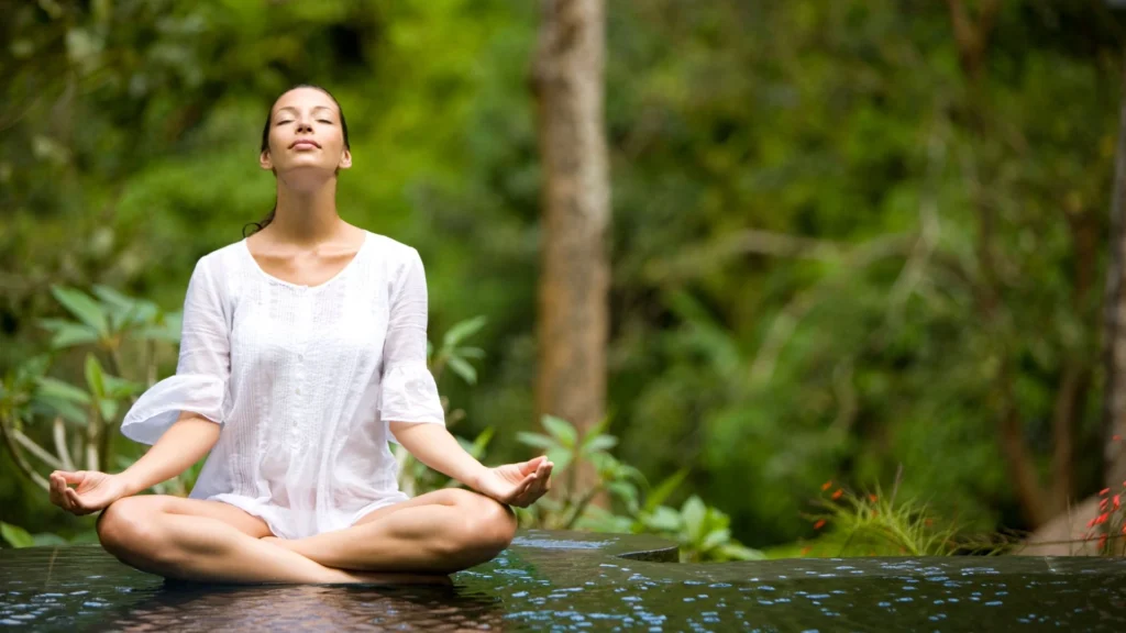 A person is doing yoga during the rainy season in Bali. The scene shows the person in a yoga pose, possibly on a covered terrace or in an indoor studio with large windows. The background is a wet, lush tropical landscape with visible rainfall.