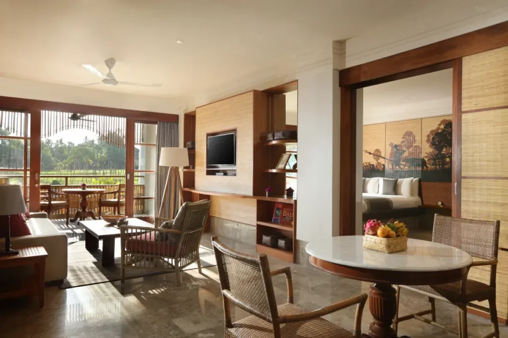 A spacious and luxurious hotel room in Bali with an opulent and classy interior design, offering a view of a rice paddy field and the rising sun from the window.