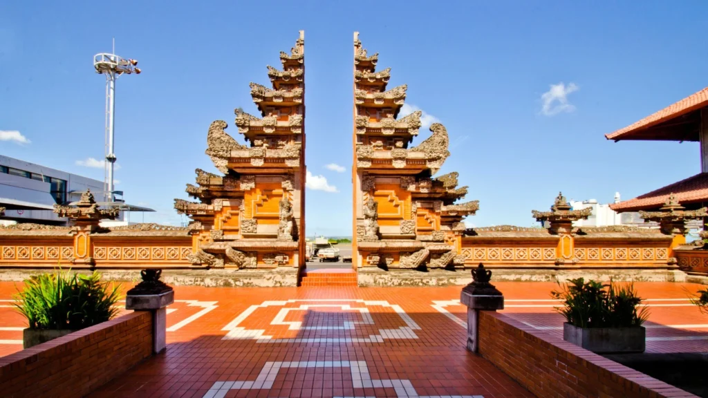 A photo of the unique exterior design of Bali's I Gusti Ngurah Rai International Airport. The building's architecture prominently features distinctive Balinese style, including traditional ornate carvings, statues, and tiered roofs.