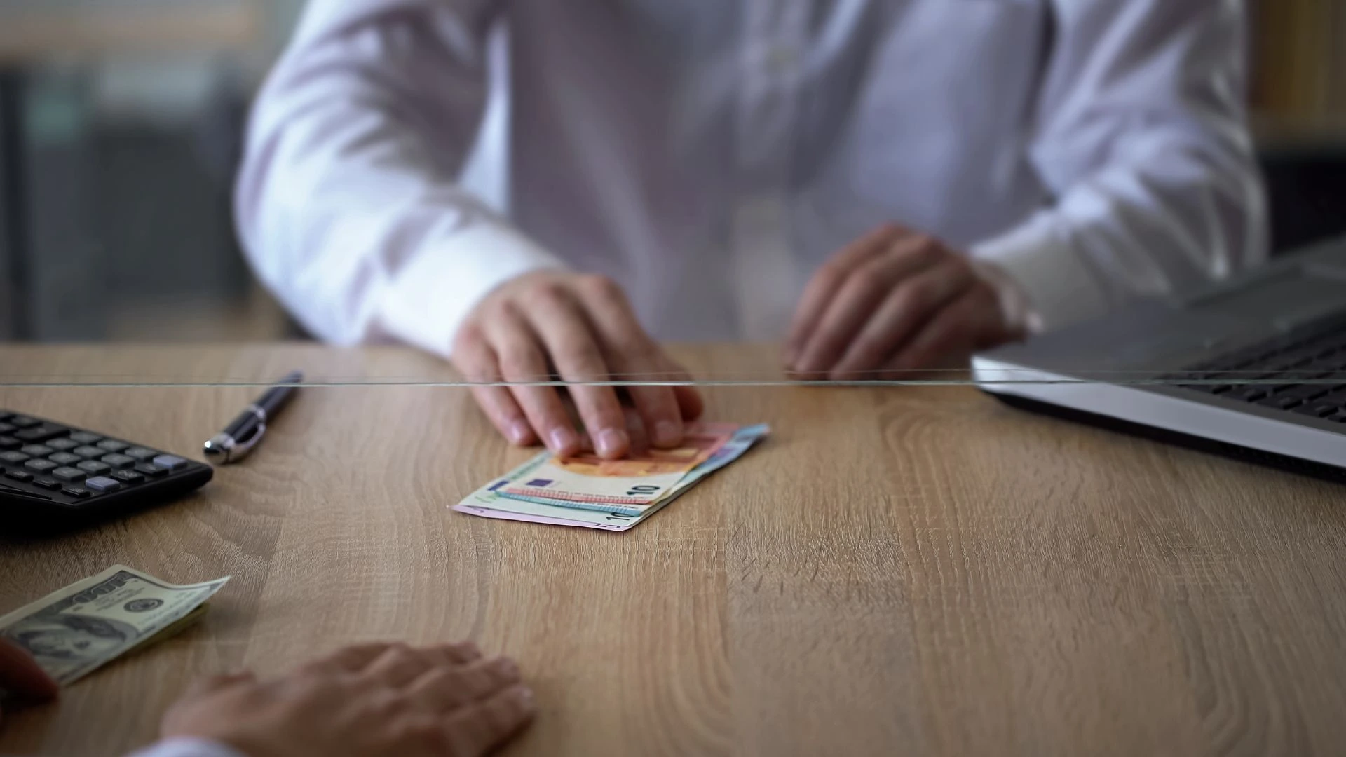 A close-up shot of a tourist at a currency exchange counter at Bali's Ngurah Rai International Airport. The image highlights a deceptive act, such as a large stack of money with a few high-denomination bills on top, while the lower ones are smaller. The tourist appears confused, representing the currency exchange scam.