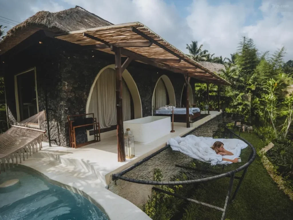 A female tourist is enjoying the natural scenery of Bali from a hammock-like net in front of her luxurious villa room. The room features a natural wooden interior design.