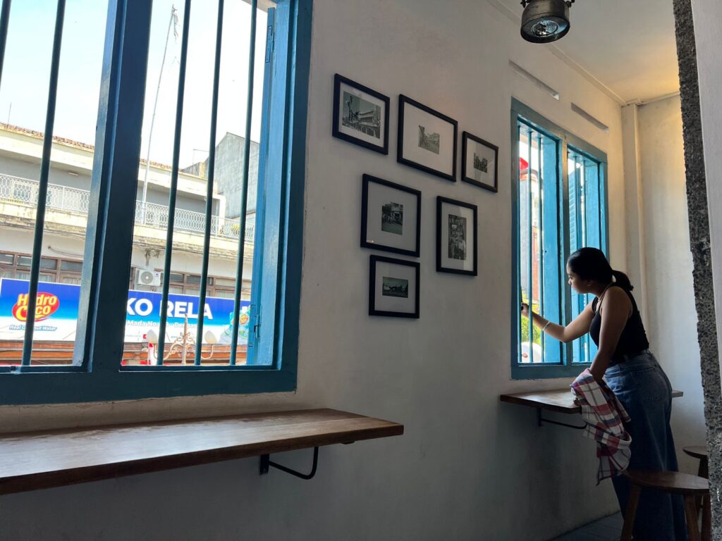 View through an upstairs window overlooking Denpasar street with honking motorbikes and people bargaining below, the scene feeling distant and nostalgic, like a glimpse of another decade.