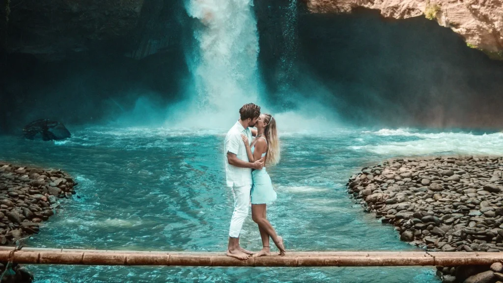 A couple is enjoying their honeymoon in Bali during the rainy season at a waterfall. They are seen smiling and embracing each other, with the lush, green tropical environment and the waterfall as a beautiful backdrop. The ground and rocks are wet, and the air looks misty from the rain and waterfall spray.