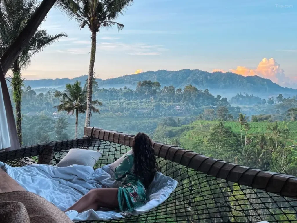 A female tourist is enjoying the natural scenery of Bali from a hammock-like net in front of her luxurious villa room. The room features a natural wooden interior design.