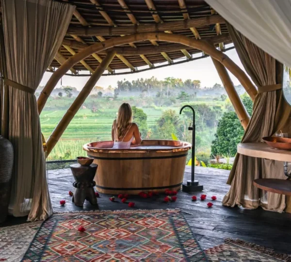 A female tourist is enjoying the natural scenery of Bali from a hammock-like net in front of her luxurious villa room. The room features a natural wooden interior design.