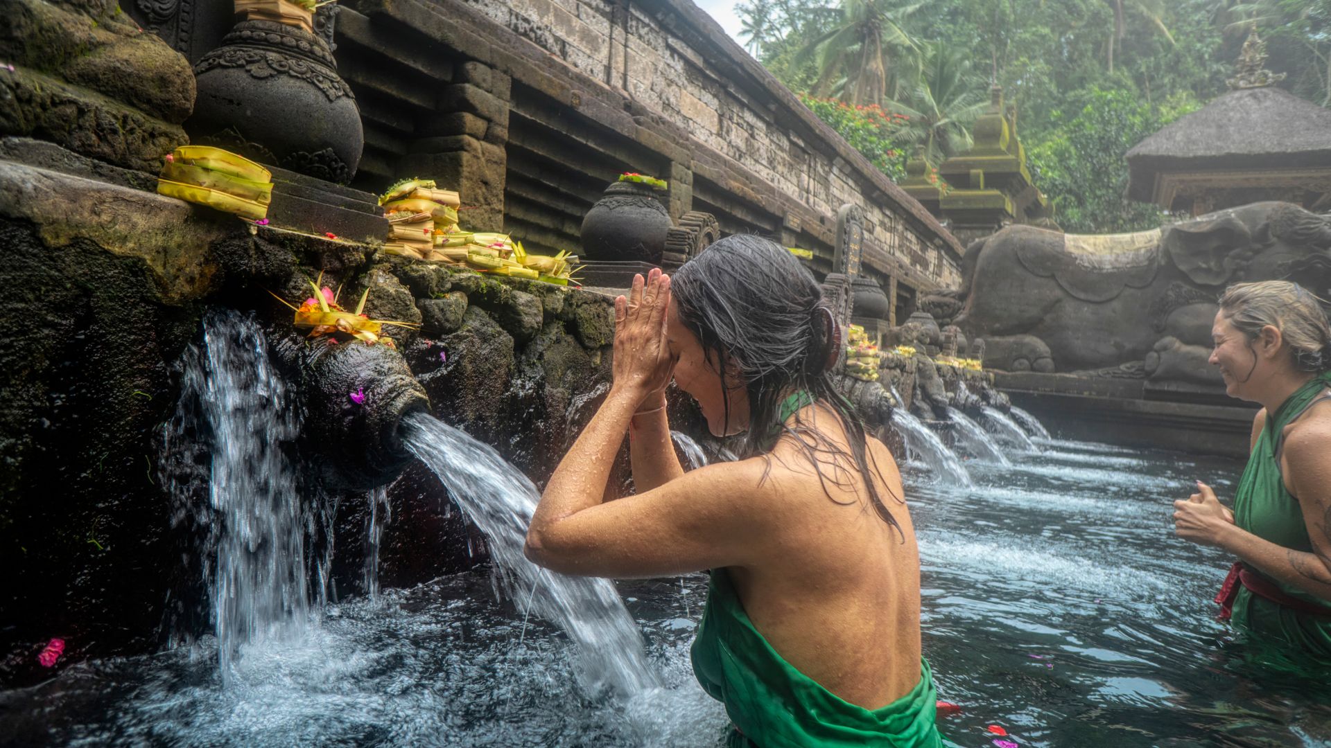A female tourist performs a melukat water purification at a spiritual site in Bali. She wears a sarong and follows the guide’s instructions with hands in prayer.