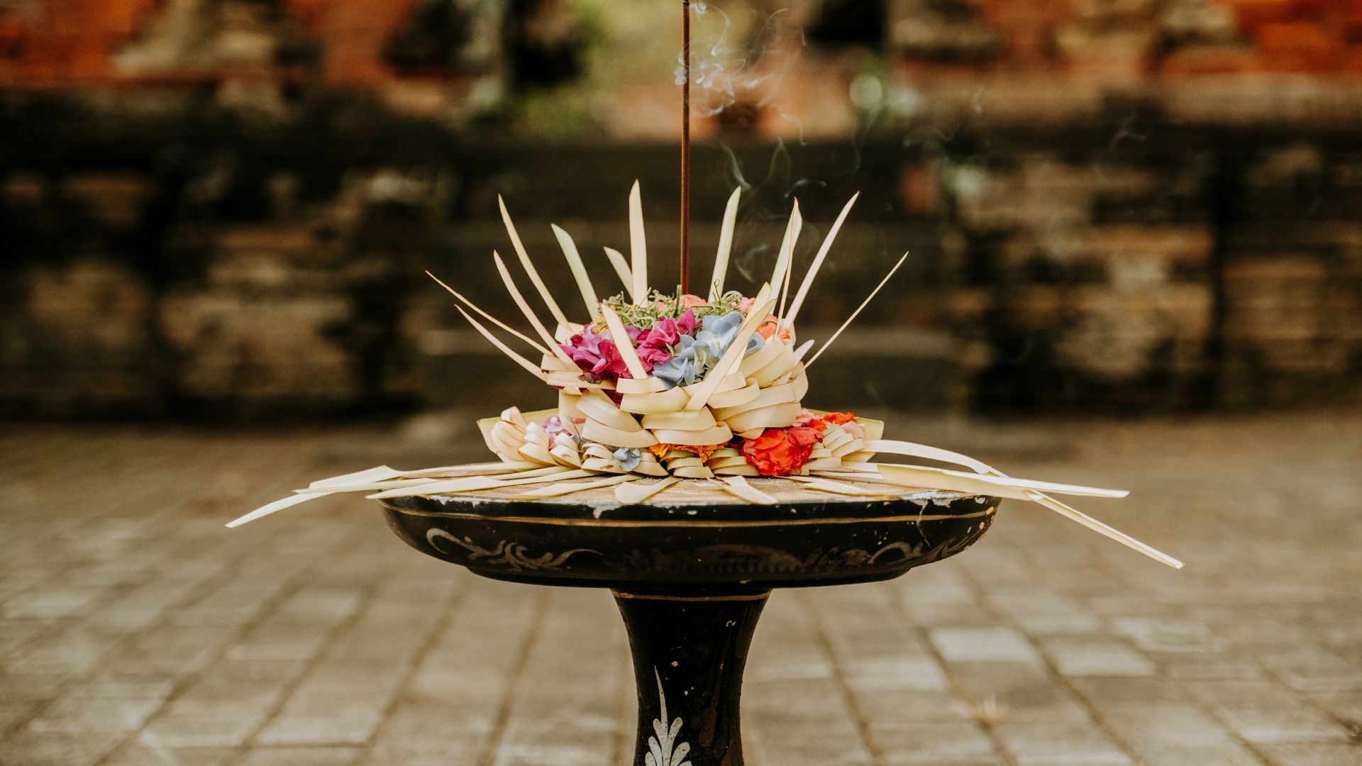 Flowers and incense sit in a small leaf tray used for Hindu Balinese prayer. Gentle smoke rises beside the colorful petals.