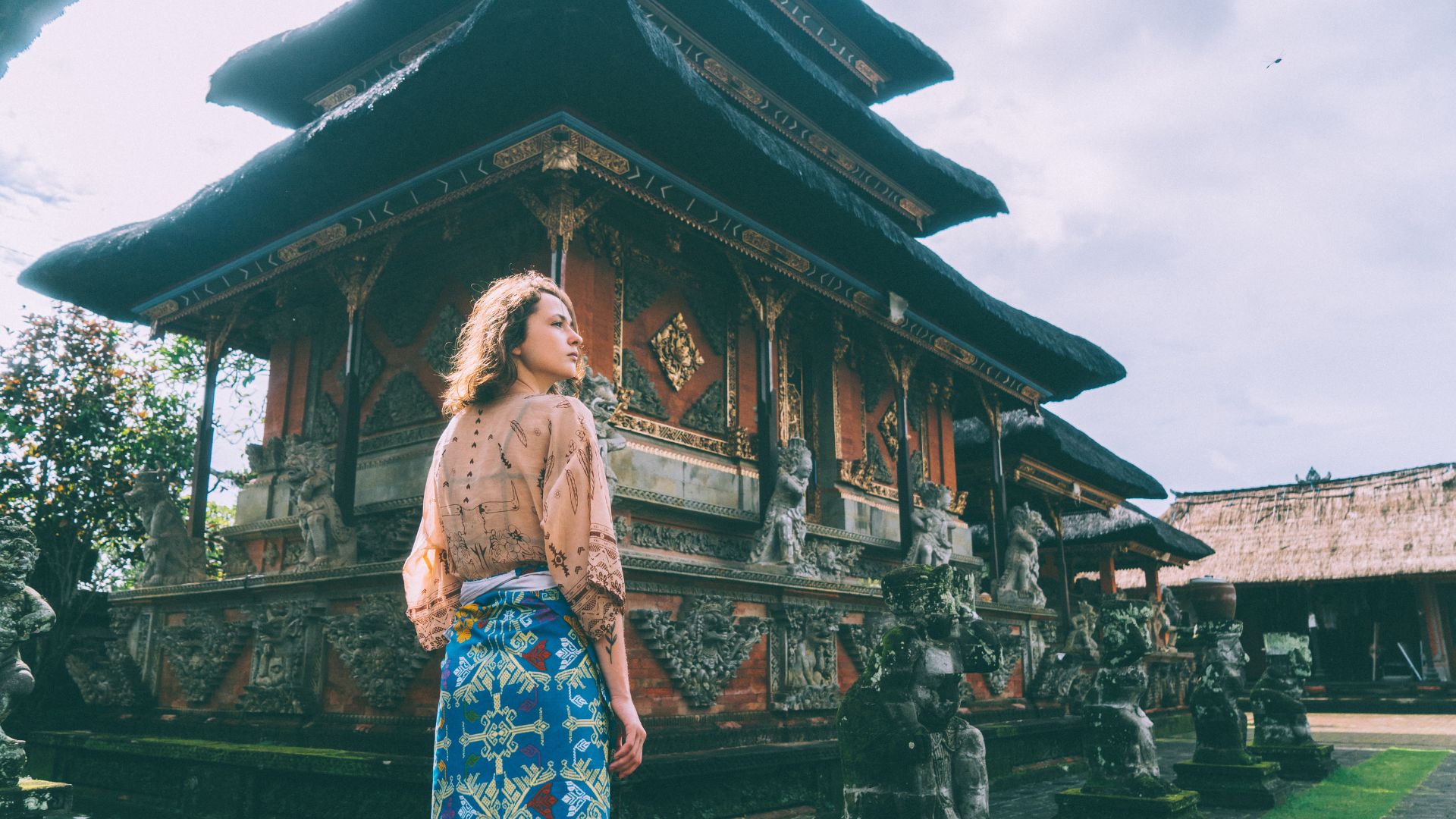 A female tourist wears modest clothing with a Balinese sarong while visiting a temple. She stands near the entrance and follows temple etiquette.