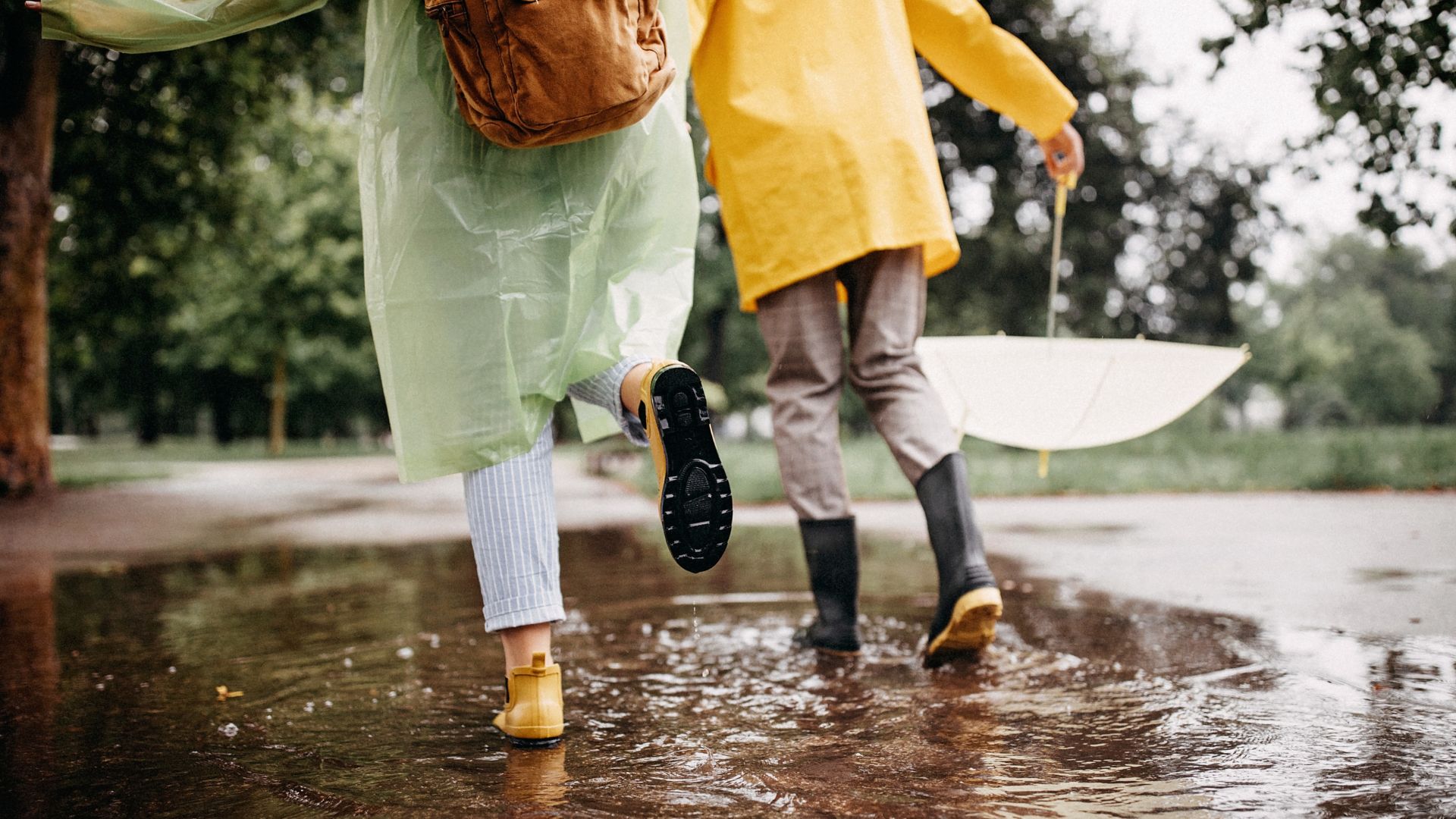A couple of tourists walk happily in the rain wearing raincoats, boots, and waterproof backpacks. They enjoy the adventure despite the wet weather.