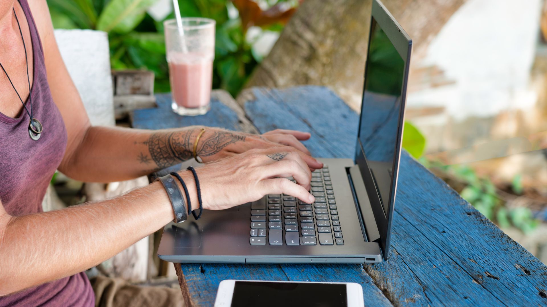 A digital nomad works at a beachside café in Bali with a natural outdoor design. The ocean is nearby and palm shade covers the table.